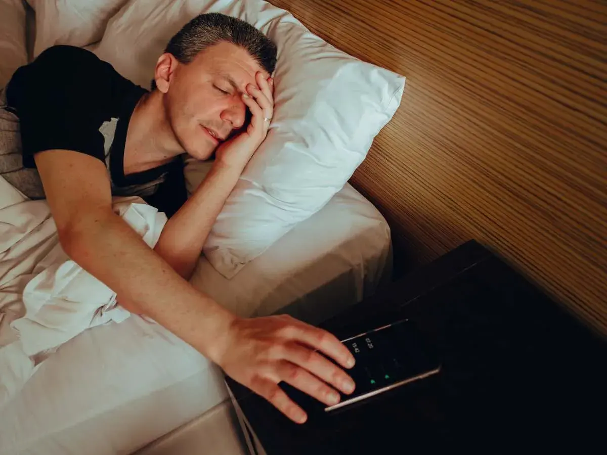 Man in bed, hand reaching for phone alarm. Does snoozing make you more tired?