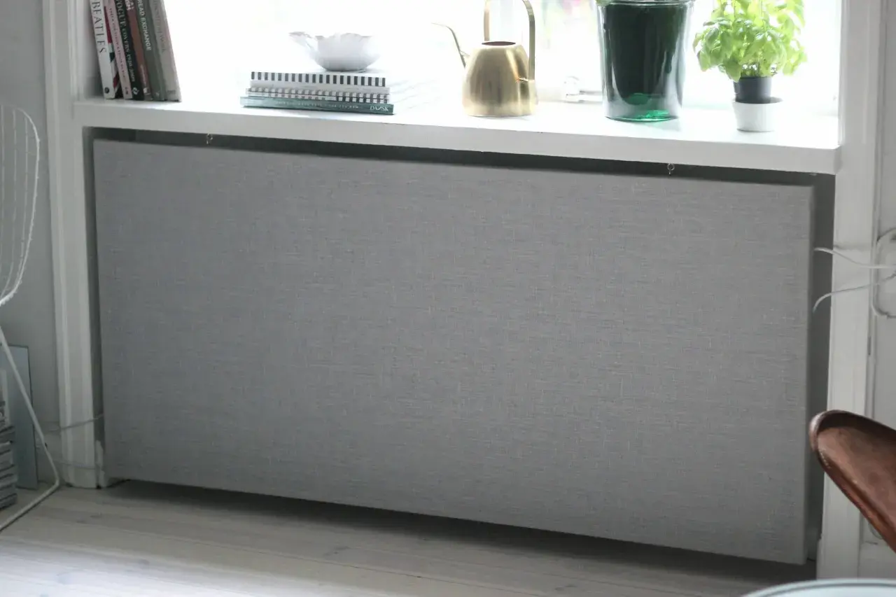 Grått elementkydd i tyg, ett DIY-projekt som ger ett stilrent intryck. Böcker och en vattenkanna står på fönsterbrädan ovanför.