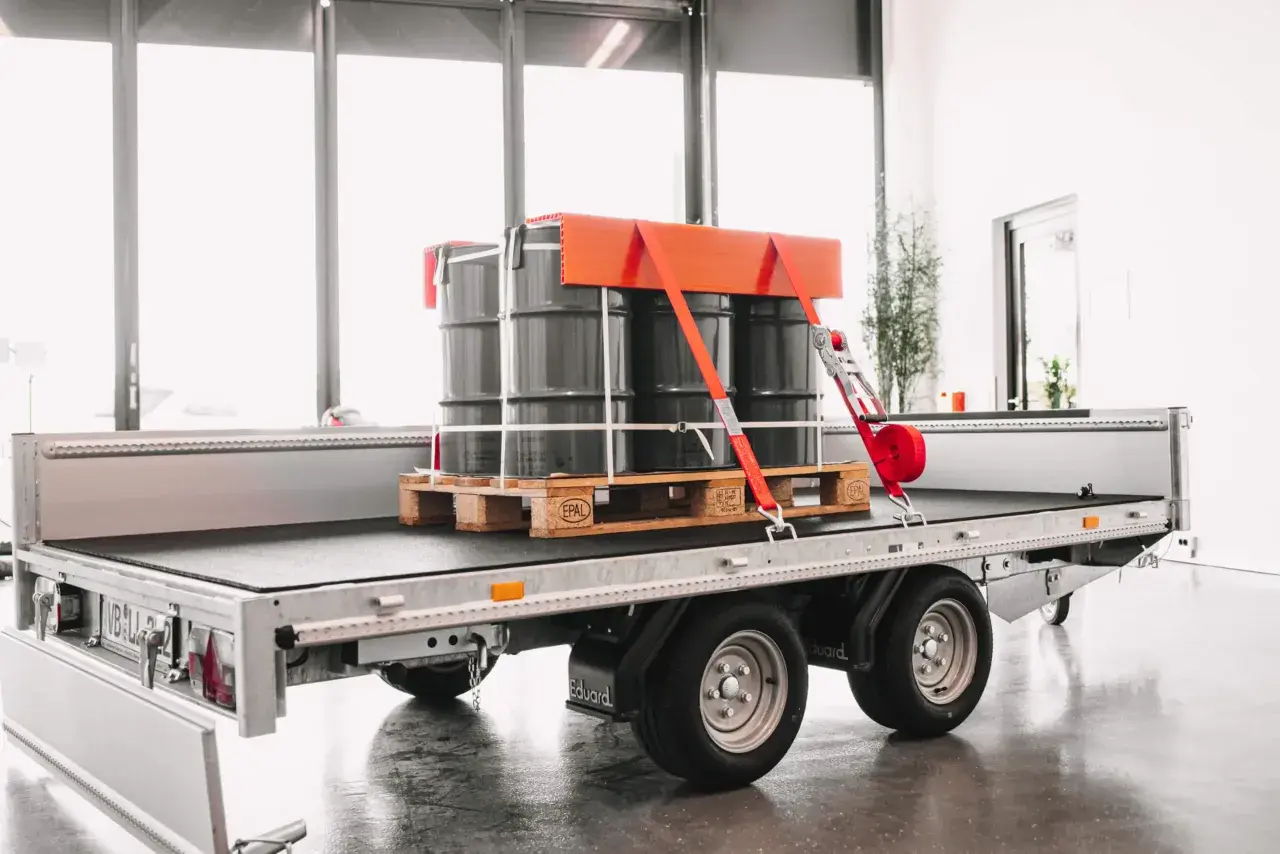 Fass auf einem Anhänger sind drei Fässer auf einer Palette sicher mit roten Gurten und einem orangefarbenen Halter frei verladen.