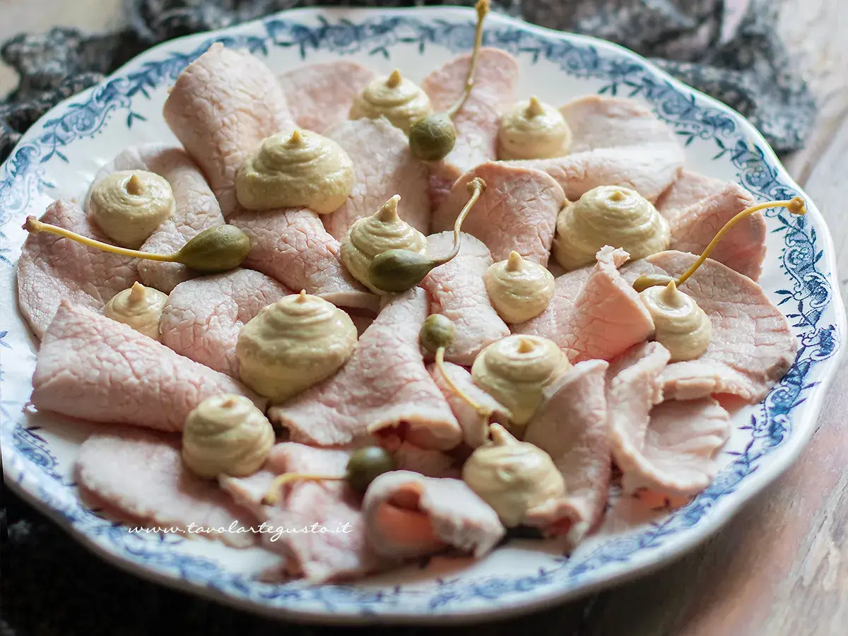 Vitello tonnato della nonna: fette sottili di carne rosa, guarnite con salsa tonnata e capperi, su un piatto bianco con decori blu.
