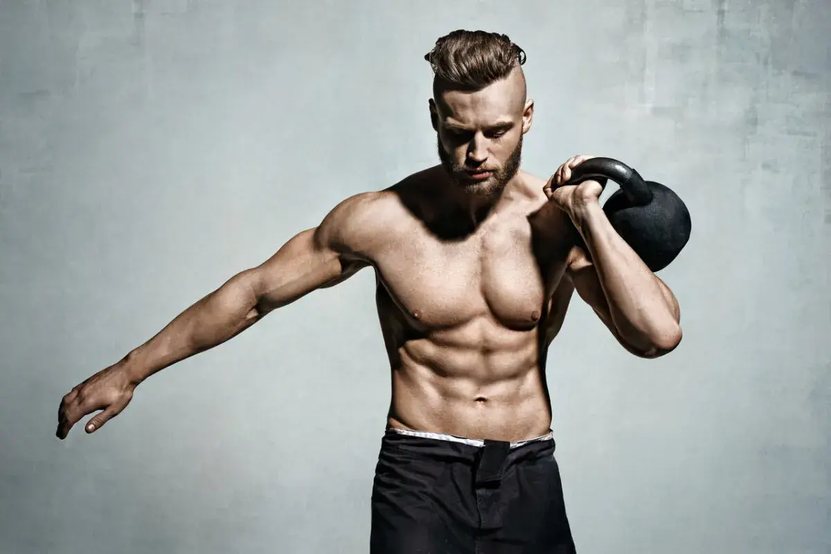 Mężczyzna z wyrzeźbionym brzuchem i kettlebell na ramieniu. Pokazuje, jak wejść na masę, trenując siłowo.