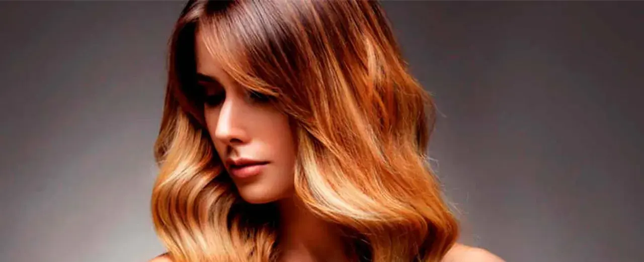 Mujer con pelo californianas, con ondas y un degradado de color castaño a rubio dorado, luciendo un estilo moderno y radiante.