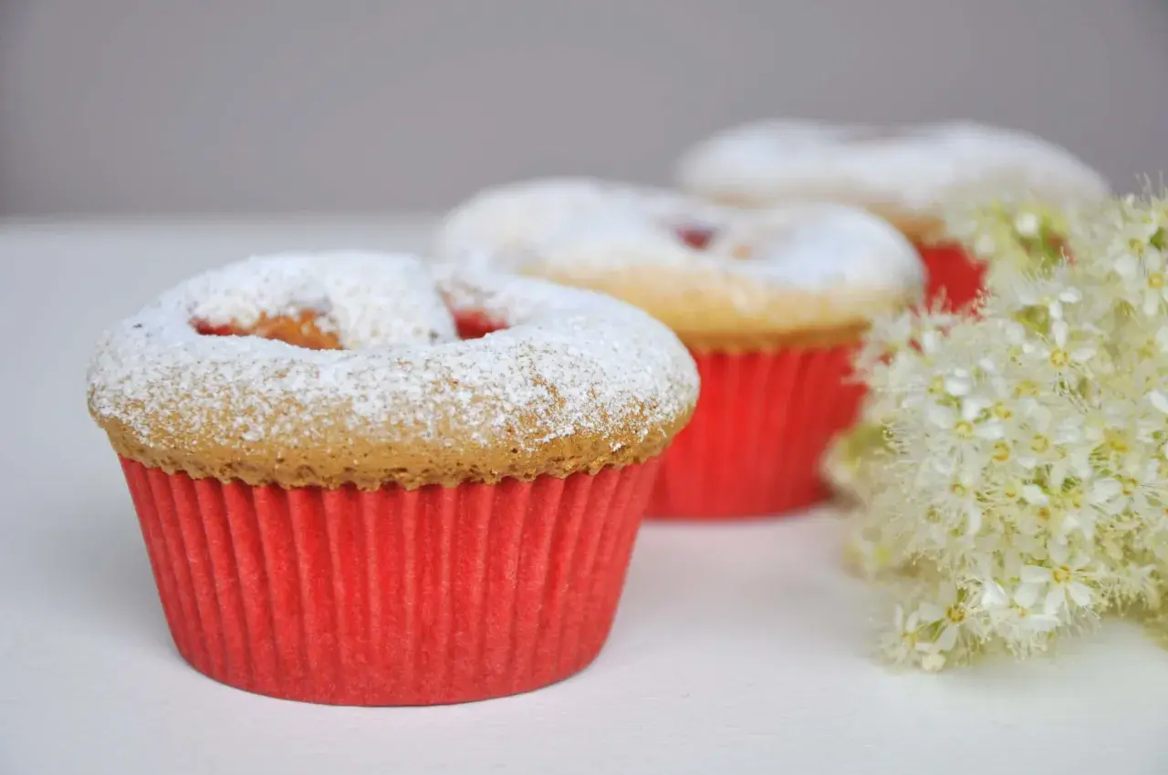 Pyszne muffinki dla dzieci, posypane cukrem pudrem, w czerwonych papilotkach, obok delikatnych białych kwiatów.
