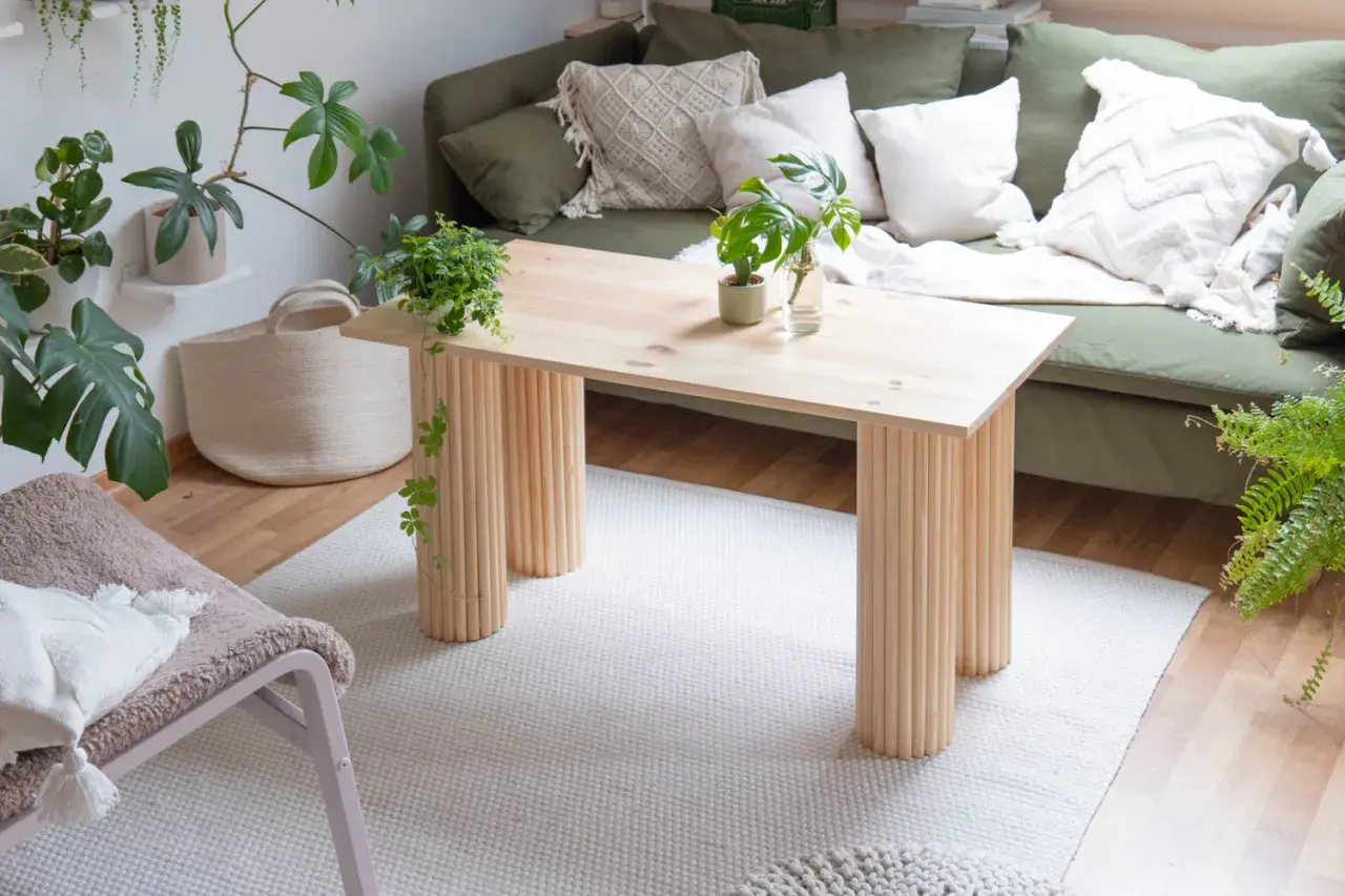 Ein heller Holztisch mit säulenartigen Beinen, perfekt für ein DIY-Projekt. Daneben ein grünes Sofa, Pflanzen und ein Körbchen.