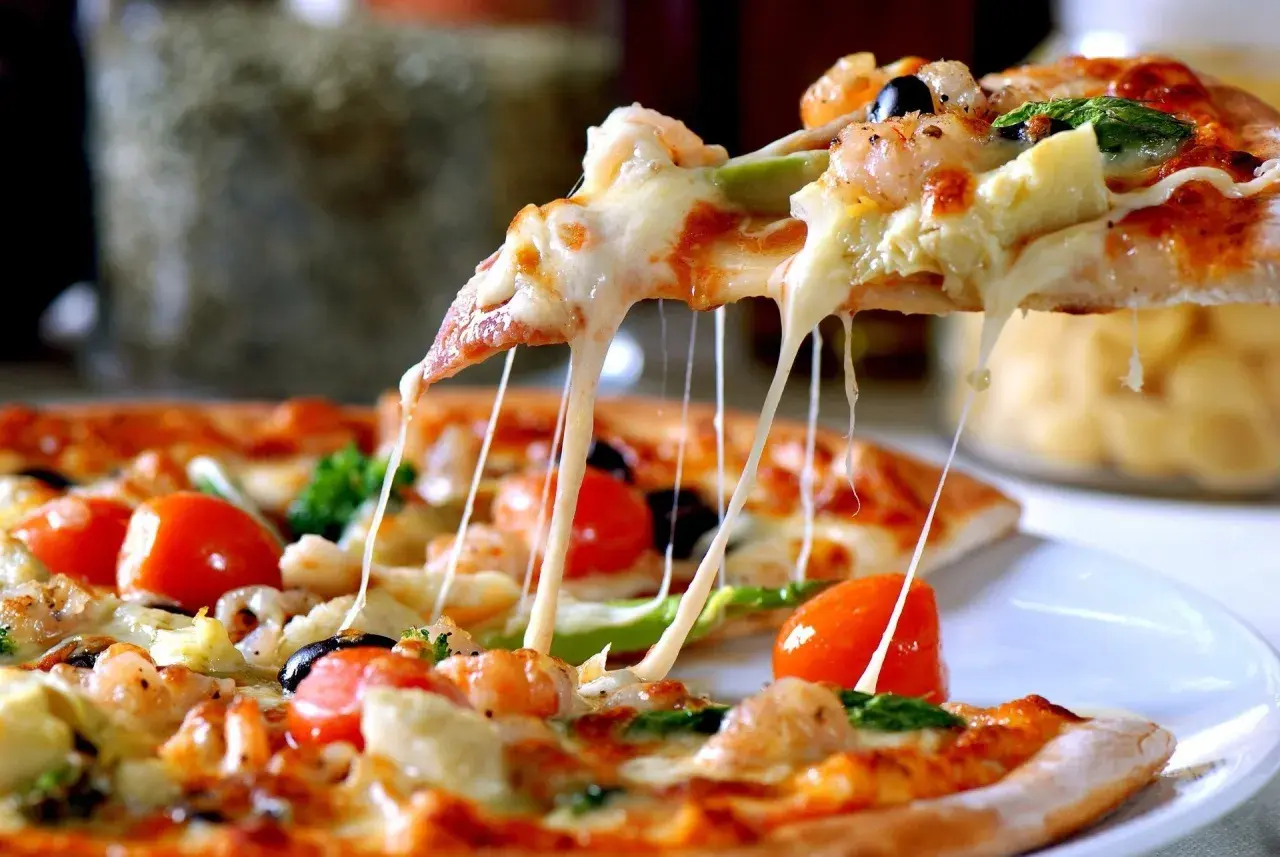 Rozciągający się ser z kawałka pizzy, na której widać krewetki, pomidorki i oliwki. Ciasto na pizzę wygląda apetycznie.