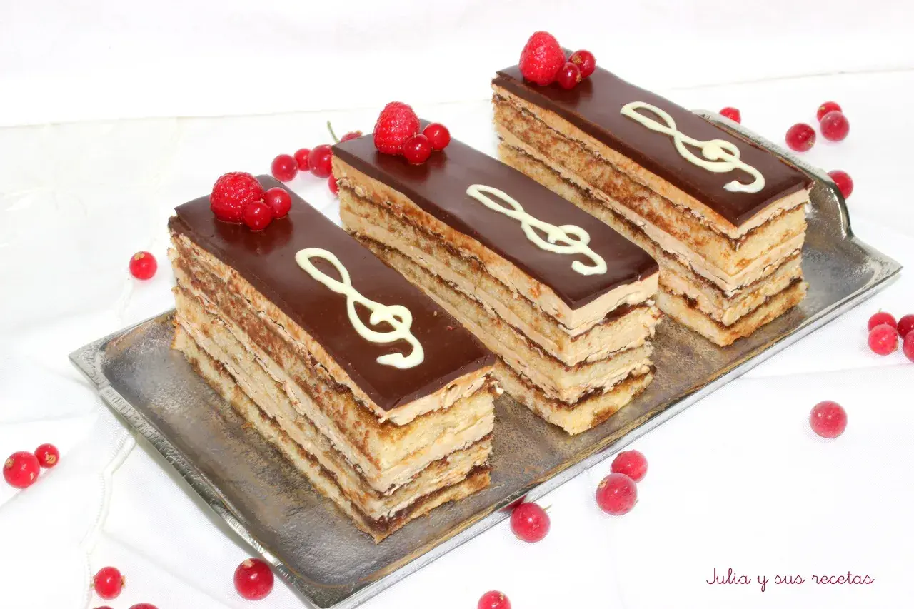 Tres porciones de pastel ópera, con capas de bizcocho y crema, cubiertas de chocolate y decoradas con clave de sol y frutos rojos.