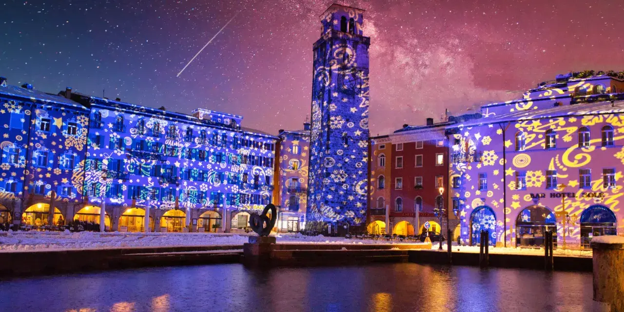 Edifici illuminati da proiezioni natalizie sul lago di Garda a dicembre, con cielo stellato e una scia luminosa.