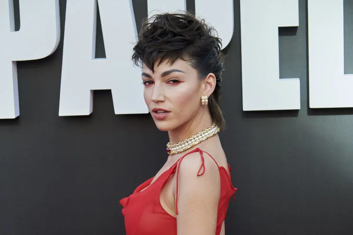 Una mujer con un peinado mullet chico y maquillaje llamativo posa con un vestido rojo y joyas doradas.