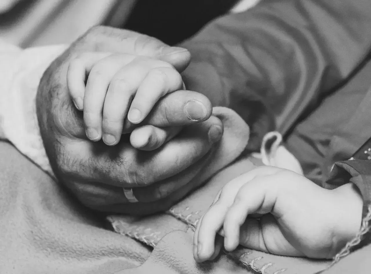 Dłoń dorosłego obejmuje maleńką dłoń dziecka. To obraz czułości i wsparcia, symbolizujący miłość rodzicielską, nawet w obliczu straty dziecka.