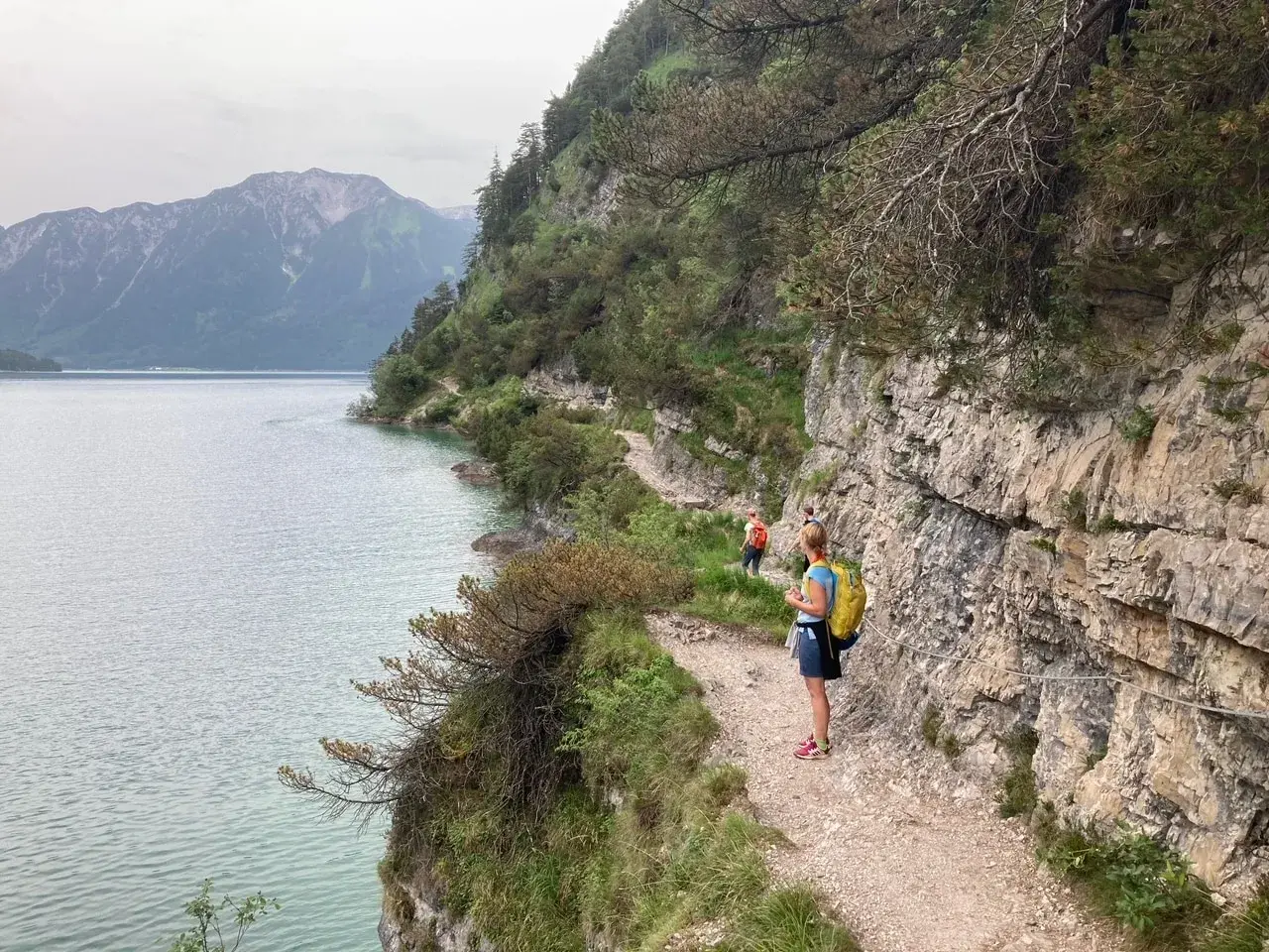 Wanderer genießen die Achensee Umrundung auf einem schmalen Pfad entlang steiler Felswände mit Blick auf den See und die Berge.