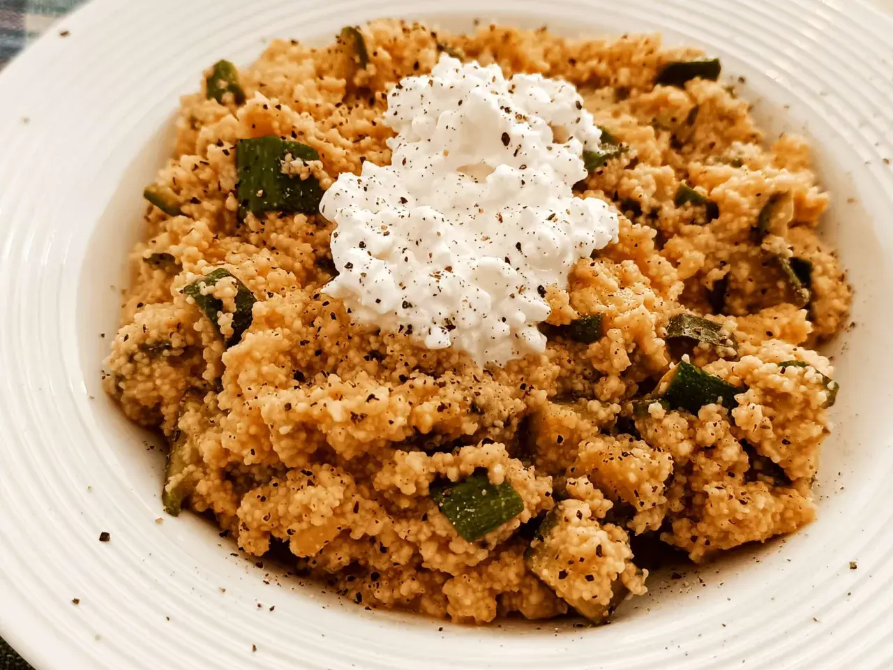 Un piatto di cous cous con zucchine e una generosa porzione di fiocchi di latte al centro, condito con pepe nero.