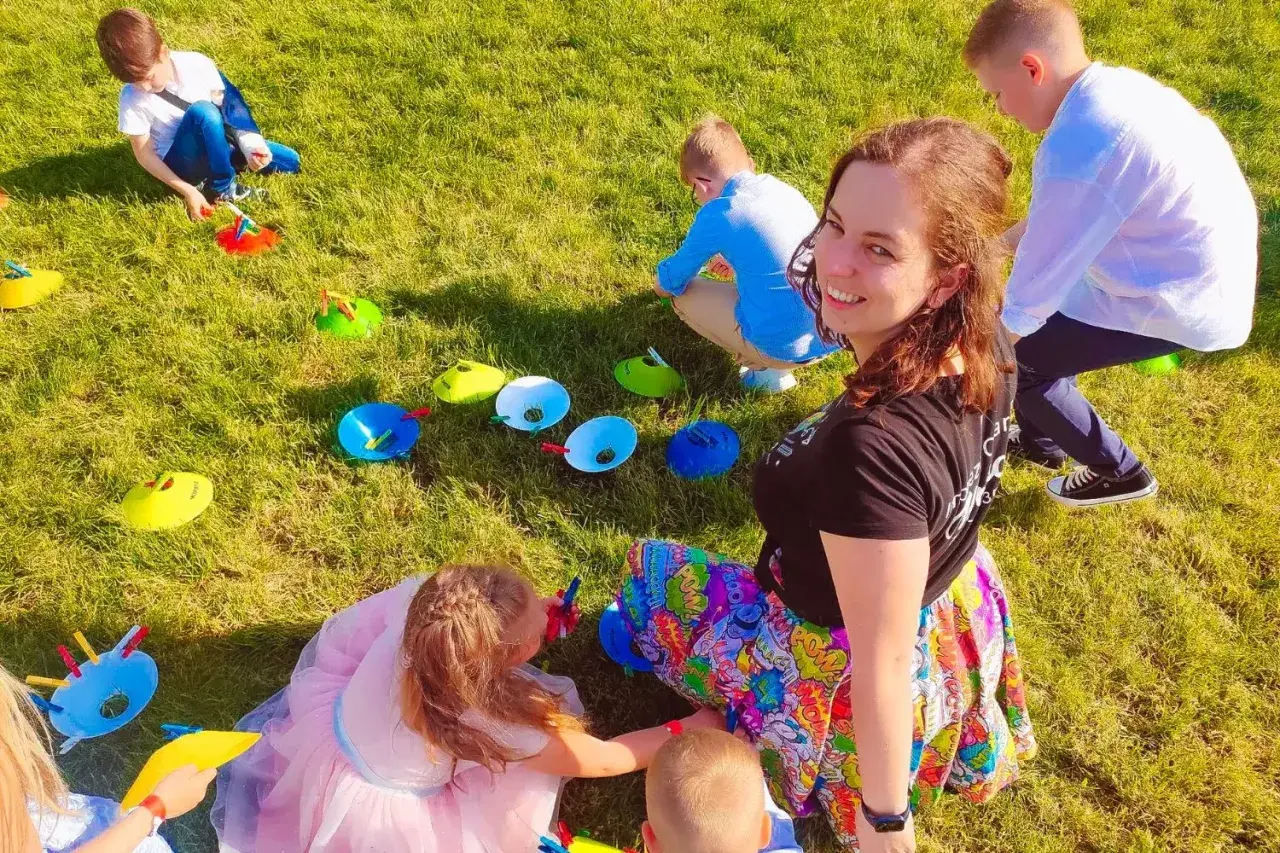 Uśmiechnięta animator dla dzieci cena. Dzieci bawią się na trawie, wykonując zadania z kolorowymi pachołkami.