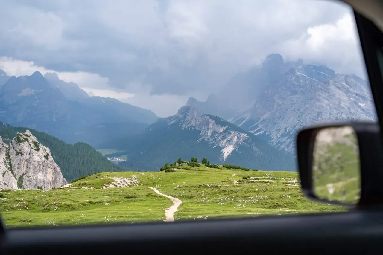 Vue depuis une voiture en Italie, traversant des prairies verdoyantes et des montagnes majestueuses sous un ciel nuageux.
