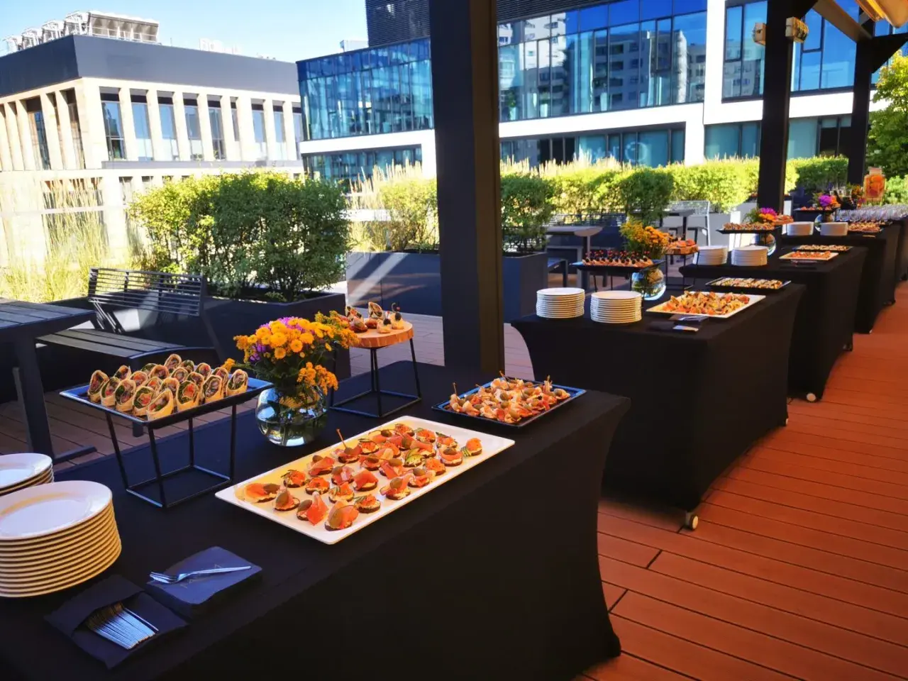 Elegancki catering Warszawa na imprezę plenerową. Stoły z przekąskami i napojami na tarasie z widokiem na nowoczesne budynki.