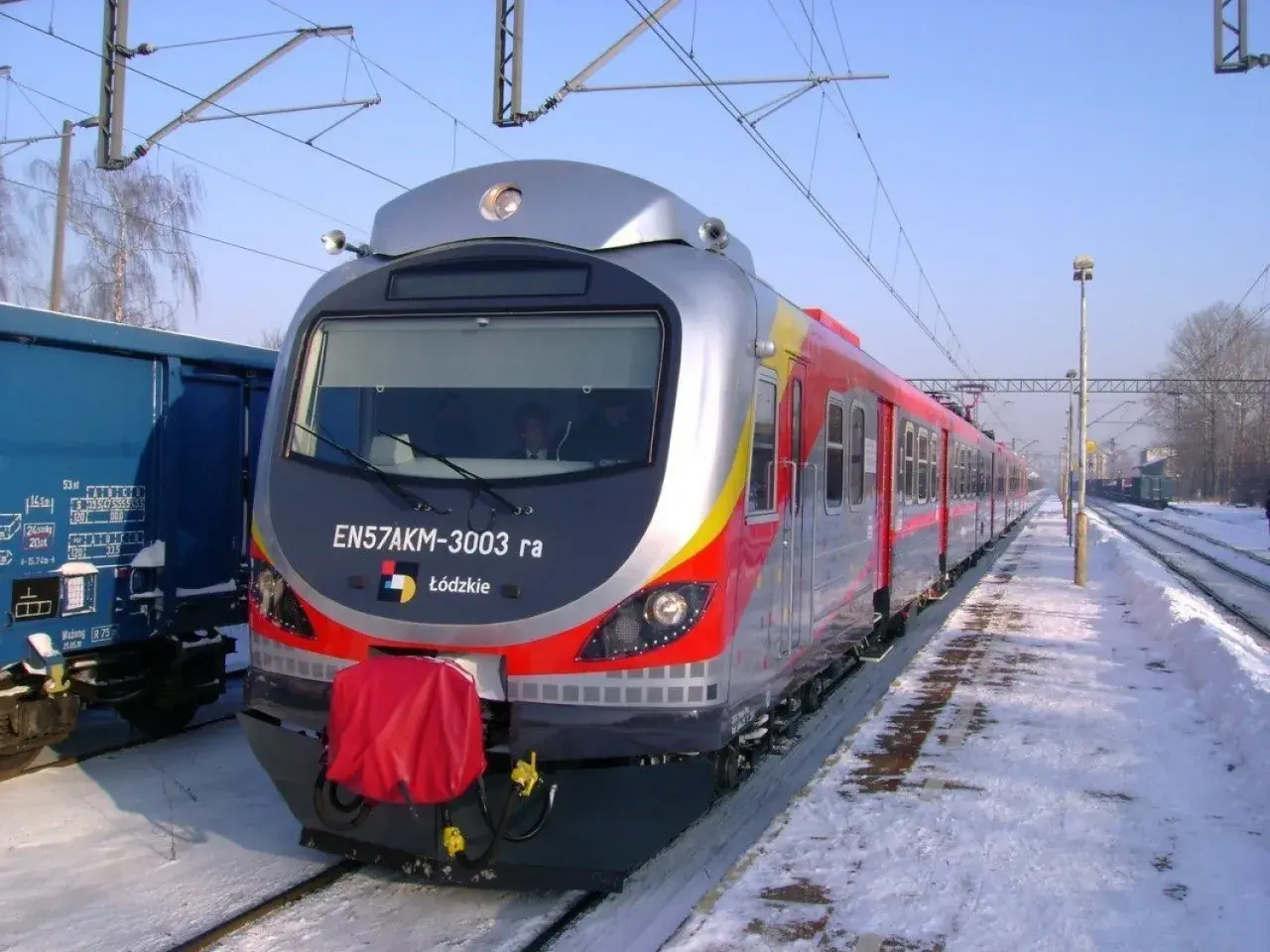 Pociąg EN57AKM-3003 ra z Łodzi stoi na zaśnieżonym peronie. Obok niebieski wagon towarowy.