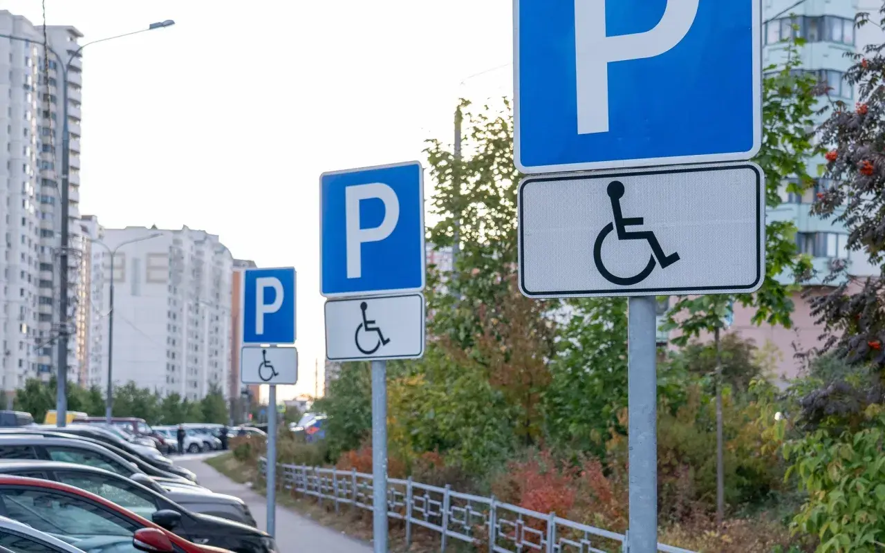Znaki informują, komu przysługuje karta parkingowa. Na parkingu stoją samochody, a w tle widać budynki mieszkalne.
