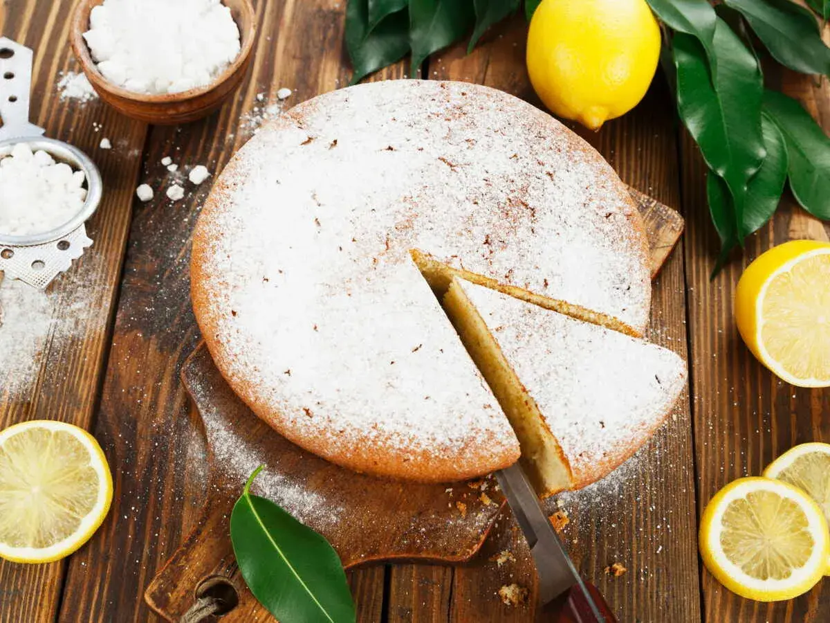 Un délicieux gâteau italien au limoncello, saupoudré de sucre glace, avec des citrons frais et des feuilles vertes sur fond de bois.