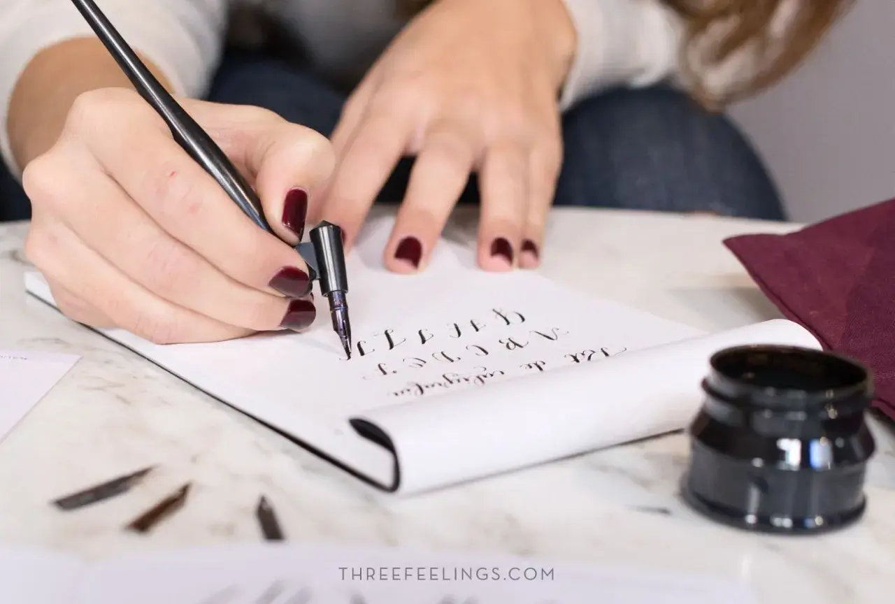 Manos practicando lettering con pluma y tinta, creando letras cursivas en un cuaderno.
