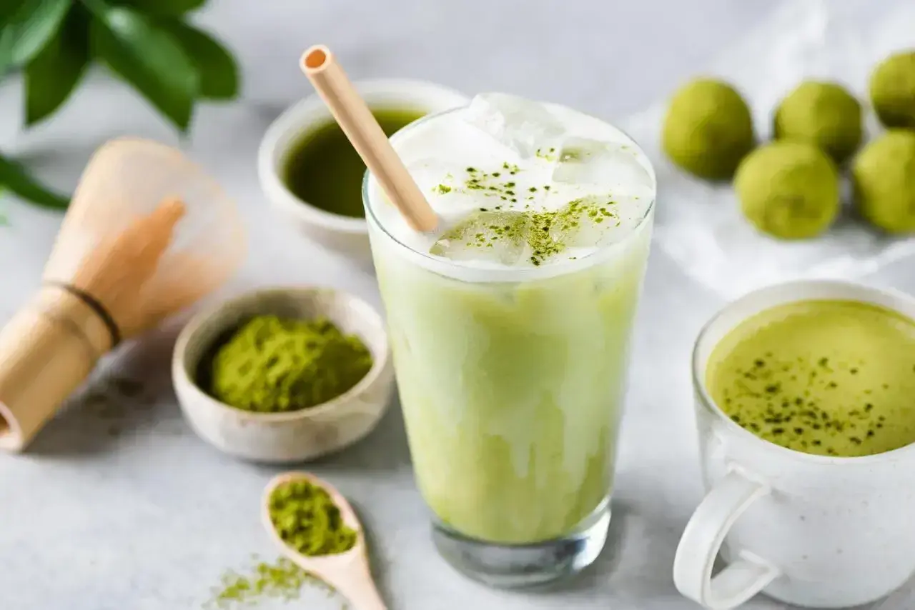 Orzeźwiający iced matcha latte z pianką i bambusową słomką, obok miseczki z matchą i tradycyjnej miotełki do jej przygotowania.