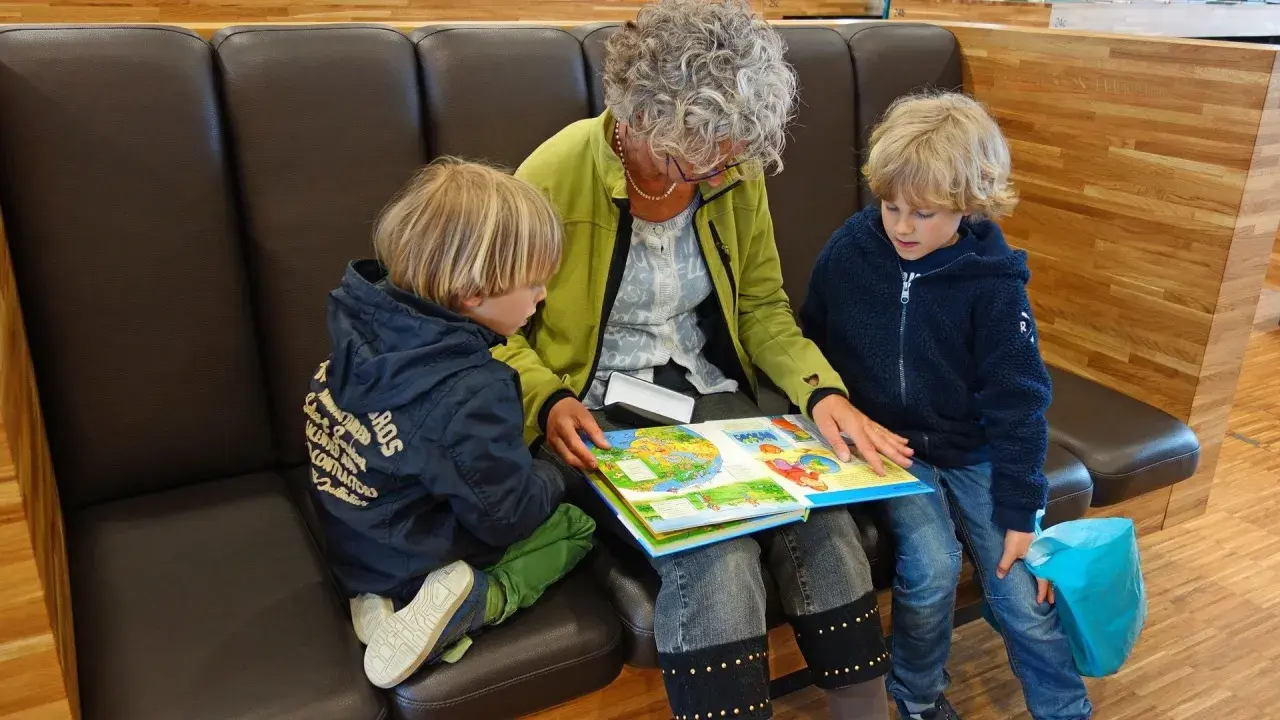 Oma und Enkel sitzen auf einer Bank und lesen gemeinsam ein buntes Bilderbuch.