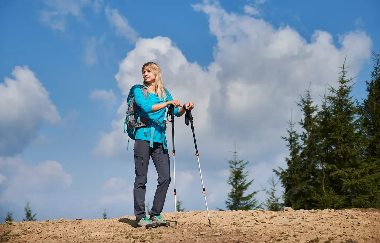 Kobieta z plecakiem i kijkami trekkingowymi na górskim szlaku. Zastanawia się, jak przymocować kijki do plecaka, by mieć wolne ręce.