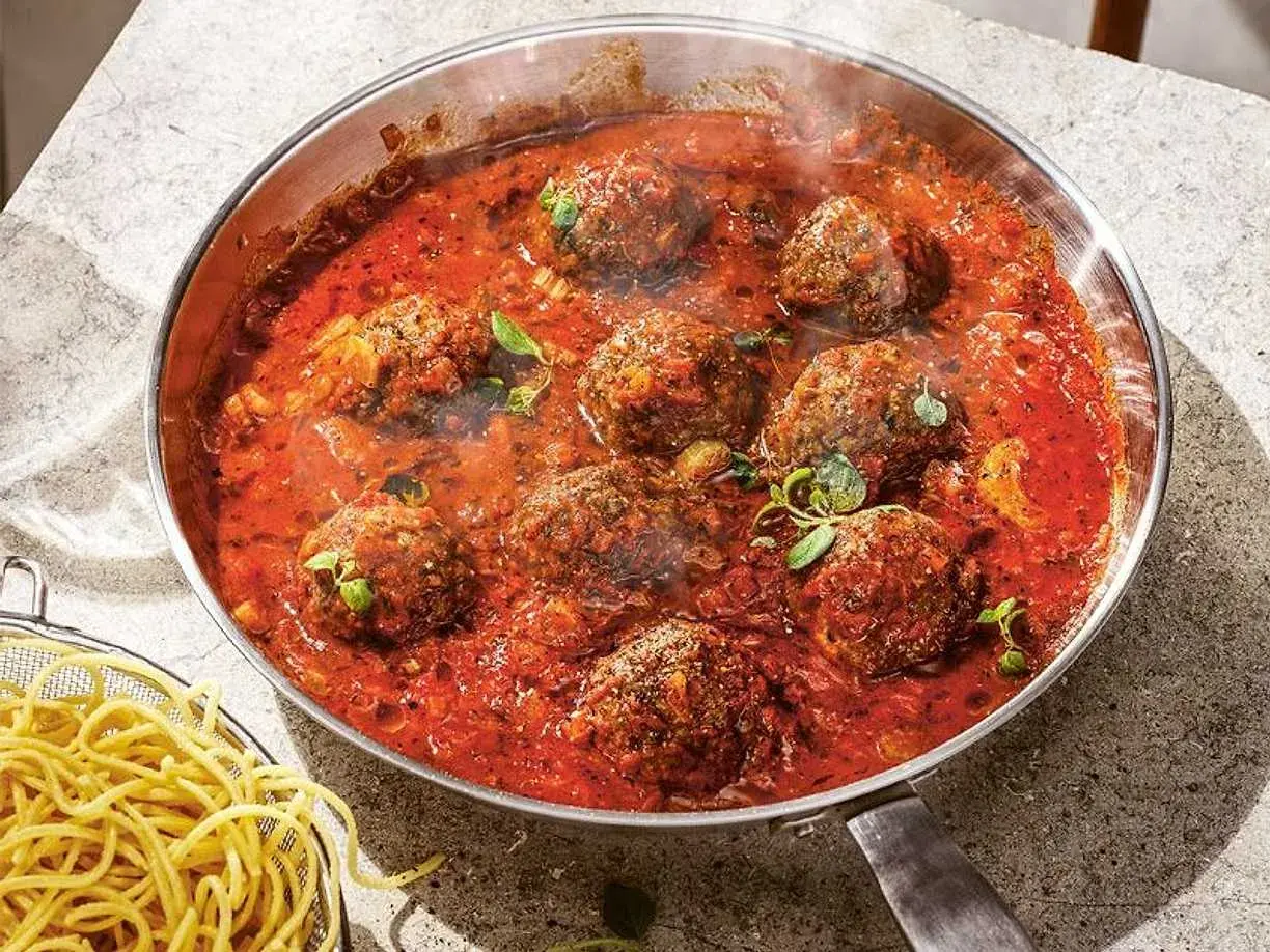 Frikadeller i tomatsås som puttrar i en panna, med en skål spaghetti bredvid.