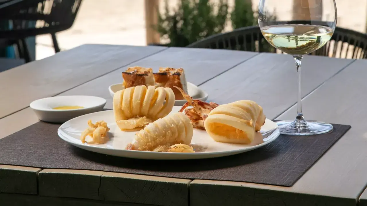 Deliciosa fritura de pescado, calamares crujientes y anillas de calamar, acompañados de pan tostado y una copa de vino blanco.