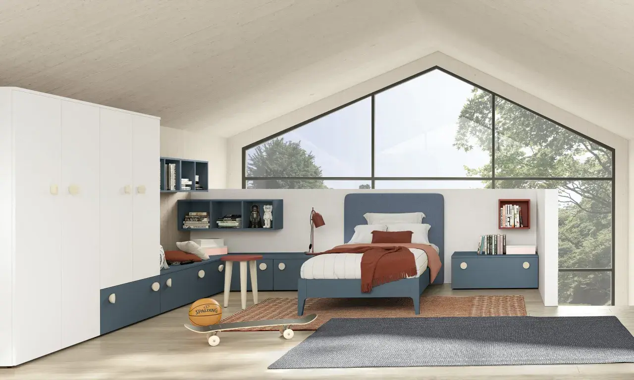 Un accogliente **letto sotto la finestra** in una stanza moderna con armadio bianco, librerie blu e dettagli sportivi come pallone e skateboard.
