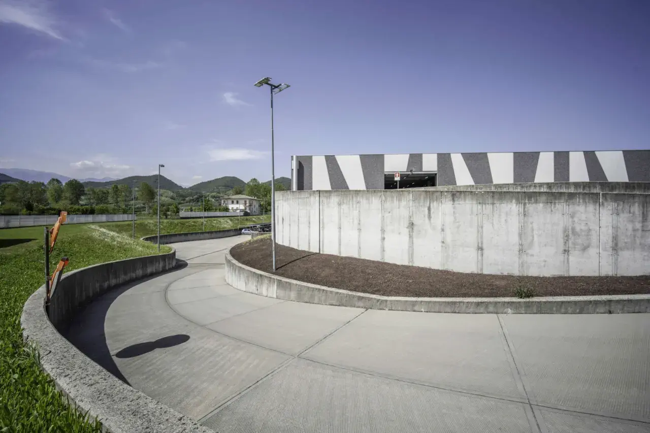 Ampia rampa in cemento fai da te, curva dolcemente verso un edificio moderno con facciata a strisce bianche e grigie, circondata da verde e colline.