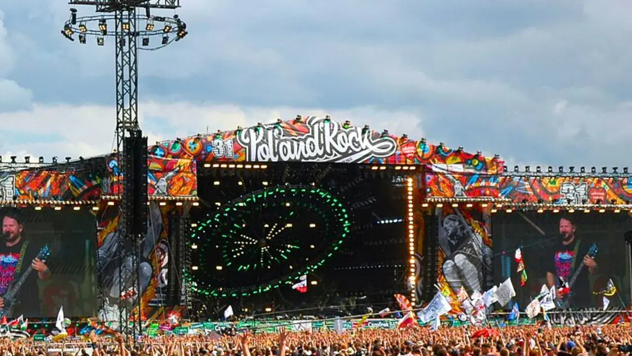 Ogromna scena festiwalowa z napisem "Poland Rock" i kolorowymi grafikami. Tłum ludzi czeka na koncert.