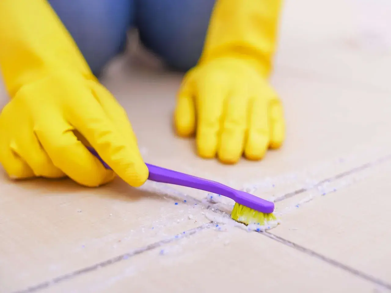 Hände in gelben Handschuhen reinigen mit einer lila Zahnbürste Fliesen und Fugen.