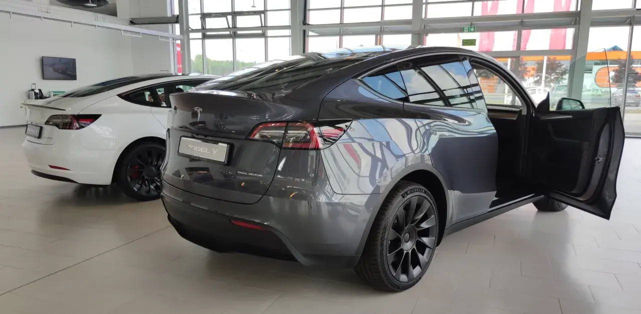 Szary Tesla Model Y z otwartymi drzwiami, obok biały Model 3. Wymiary tego SUV-a są idealne do miasta i na dalsze trasy.