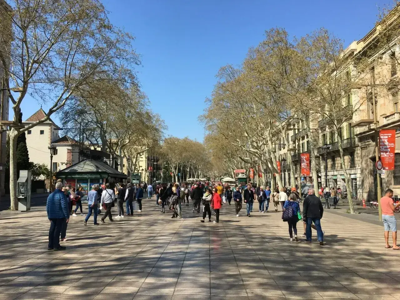 Tłum ludzi spaceruje po słynnej alei La Rambla w Barcelonie, otoczonej drzewami i zabytkowymi budynkami.