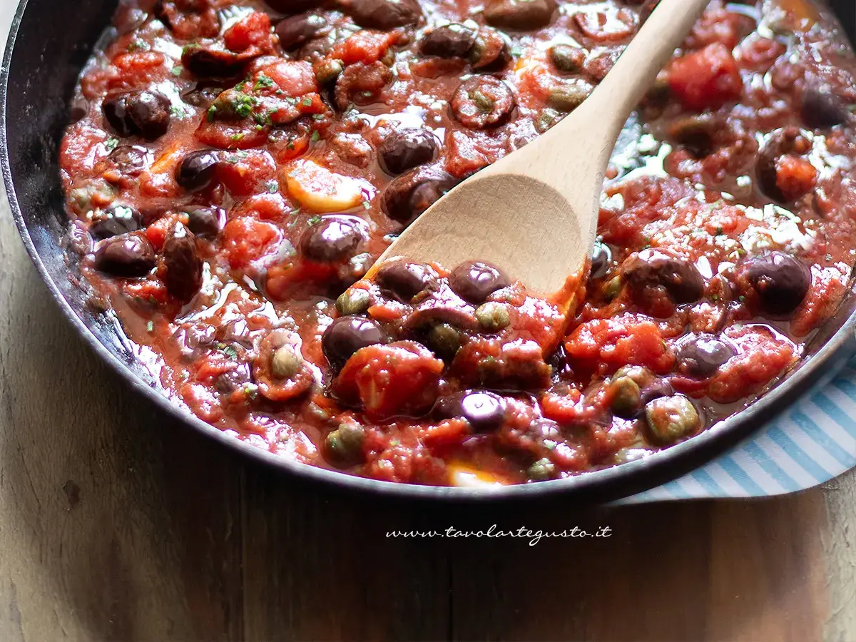 Un cucchiaio di legno mescola un sugo ricco e saporito, pieno di pomodori, olive nere e capperi. È la base perfetta per una pasta alla puttanesca.