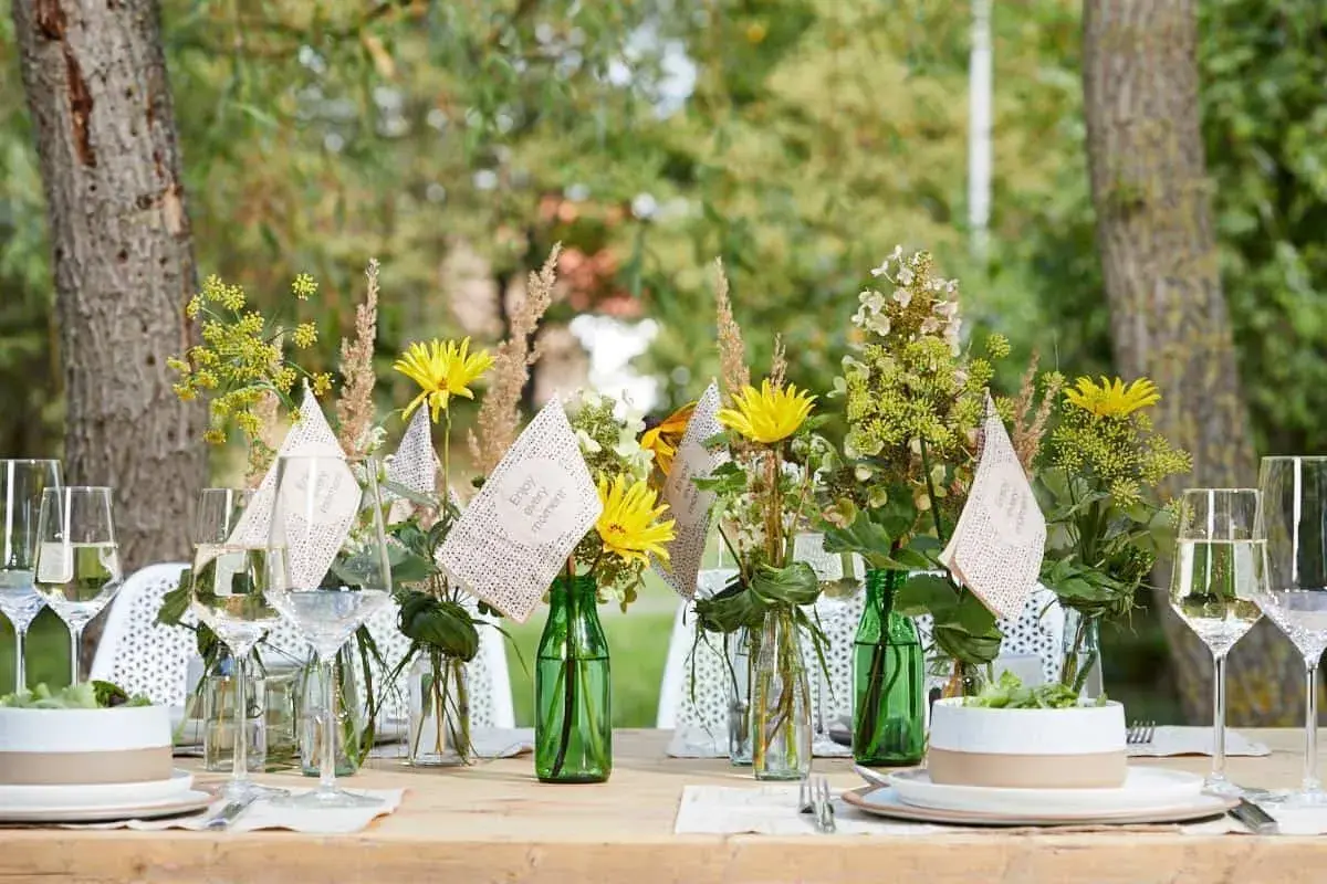 Einfache Tischdeko Sommer: Gelbe Blumen in grünen Flaschen, Weingläser und kleine Fähnchen mit der Aufschrift "Enjoy every moment" auf einem Holztisch im Freien.