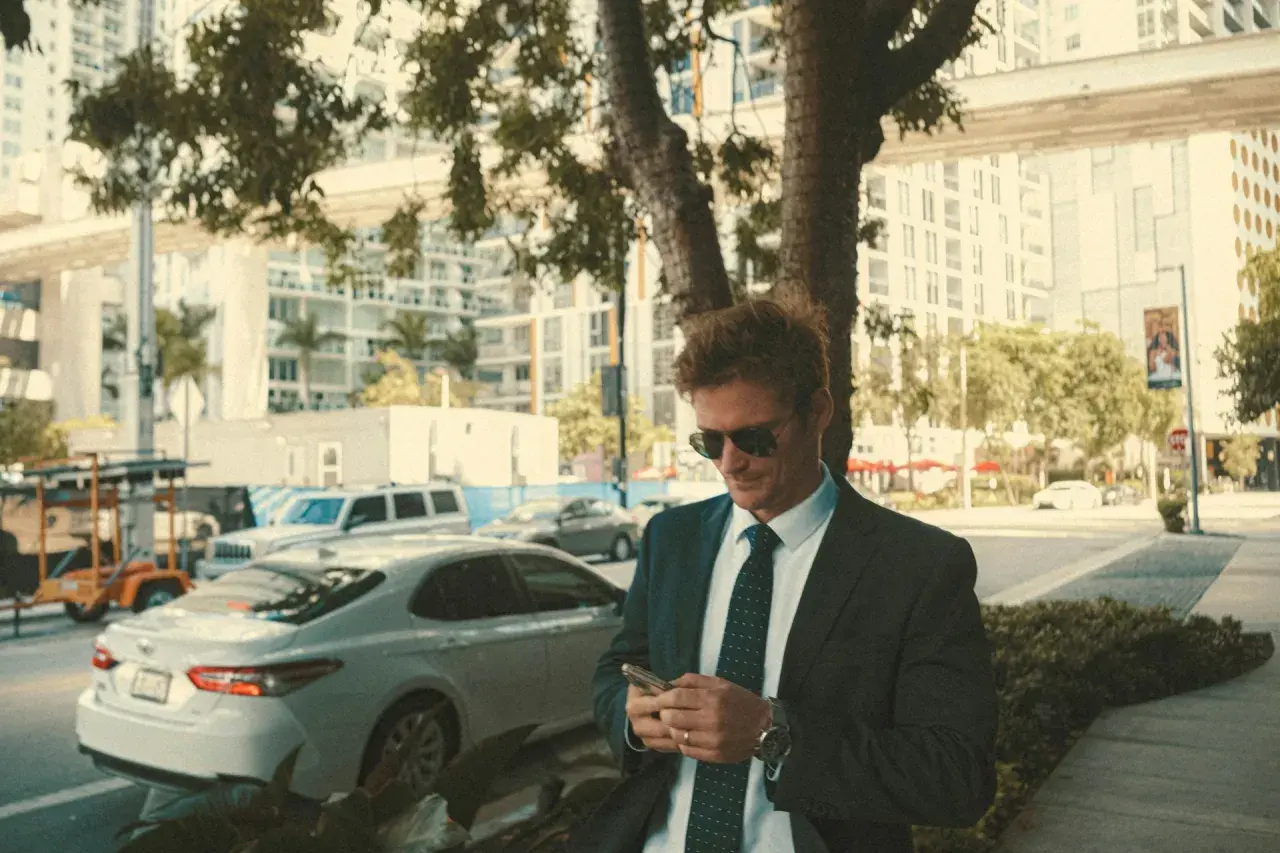 Mann im smart business dresscode, mit Sonnenbrille, blickt auf sein Handy. Im Hintergrund Stadtkulisse mit Autos.