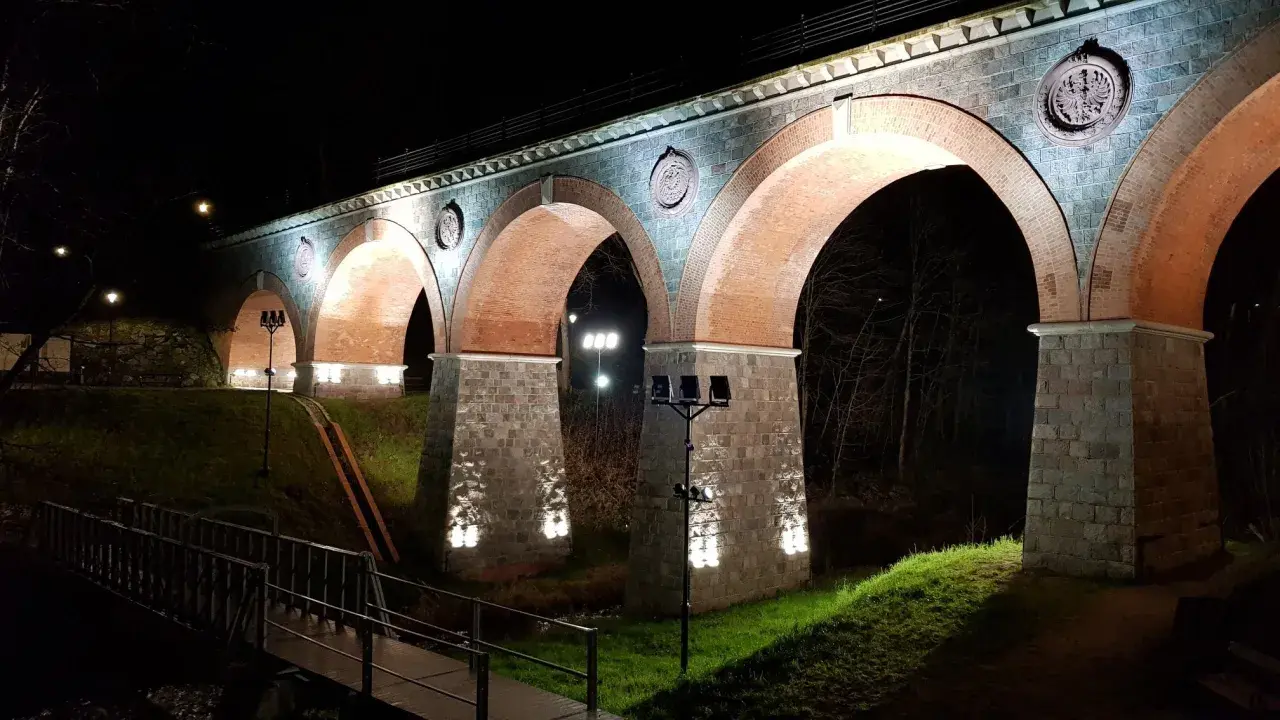 Nocna iluminacja kamiennego wiaduktu z ceglanymi łukami. Widać fragment mostku i drzewa. To piękna bytów trasa.