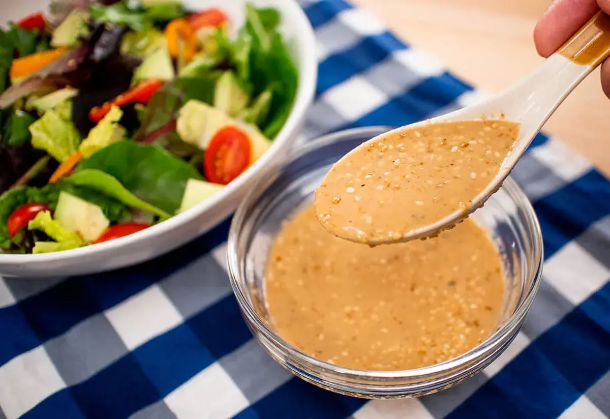 A spoon lifts creamy roasted sesame dressing from a bowl, ready to dress a fresh salad with avocado and tomatoes.