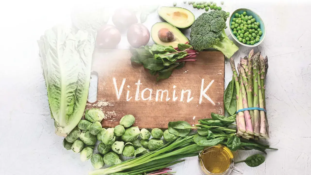 Grünes Gemüse wie Spinat, Brokkoli und Rosenkohl, das auf einem Holzbrett mit der Aufschrift "Vitamin K" arrangiert ist, zeigt, was Vitamin K ist.