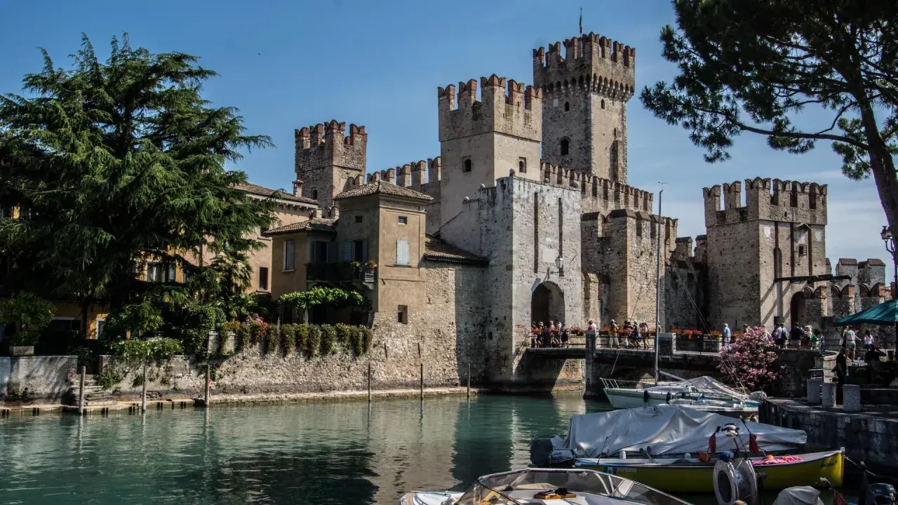 Il castello di Moniga si affaccia sull'acqua, con torri merlate e un ponte pedonale affollato. Barche ormeggiate completano la scena.