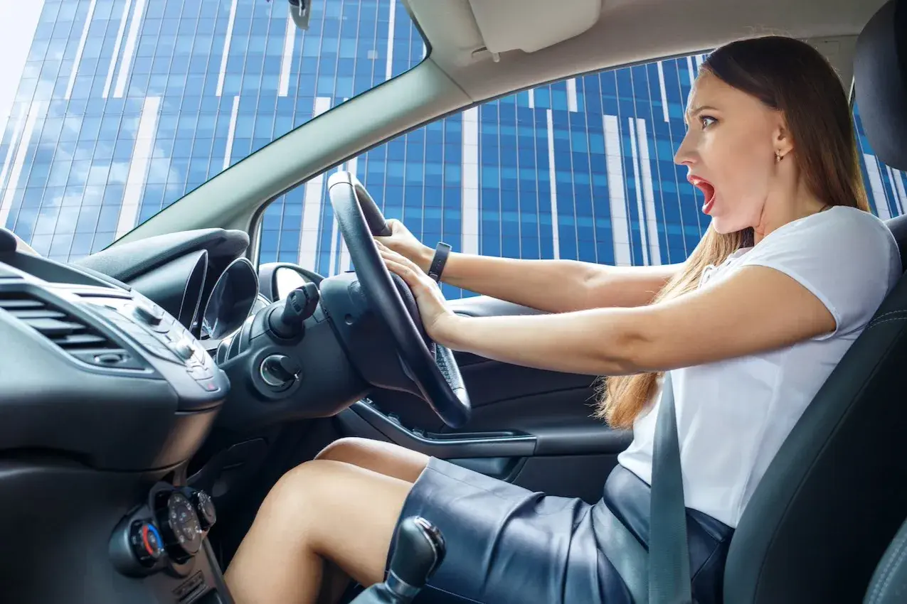 Mujer con expresión de susto al volante, parece que escucha un chirrido al girar el volante mientras conduce frente a un edificio moderno.