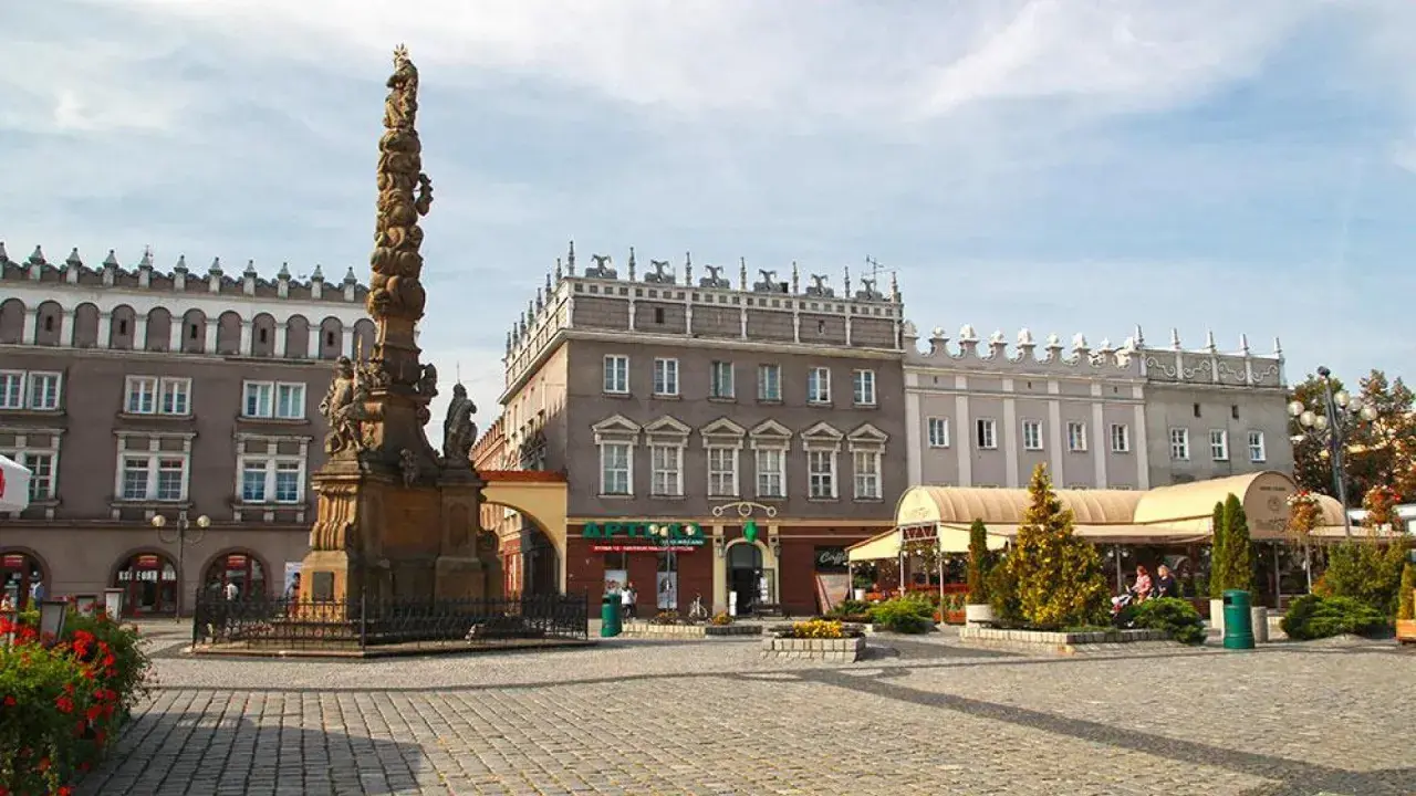 Rynek w Raciborzu z zabytkową kolumną i kamienicami. W tle apteka i kawiarnie z ogródkami.
