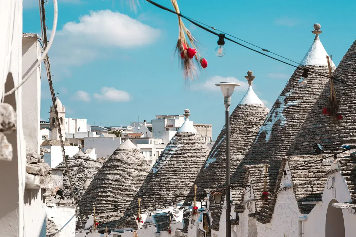 Scorci di Alberobello con i caratteristici trulli, alcuni decorati con simboli bianchi, sotto un cielo azzurro. Una scalinata cuori conduce tra le abitazioni uniche.