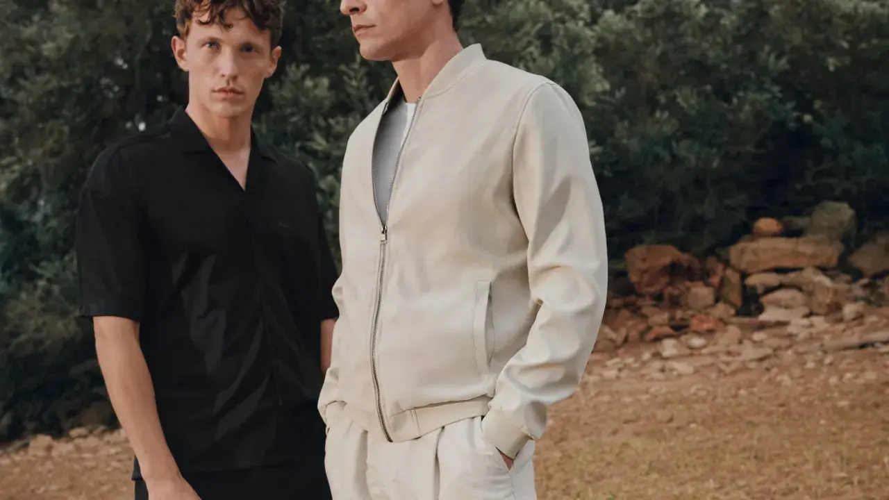 Zwei Männer im lässigen Party-Chic-Look: Einer trägt ein schwarzes Hemd, der andere eine cremefarbene Bomberjacke.