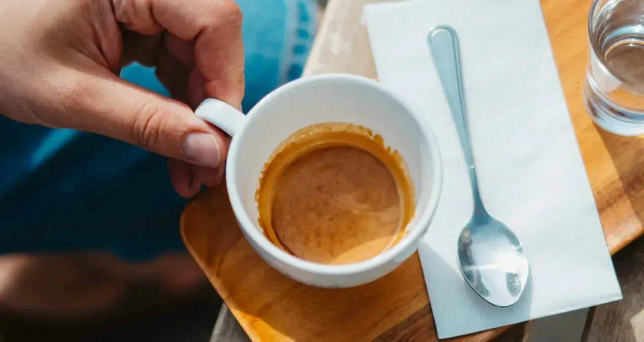 Dłoń trzyma filiżankę z espresso. Idealna ilość kawy na espresso to około 7-9 gramów. Obok leży łyżeczka i szklanka wody.