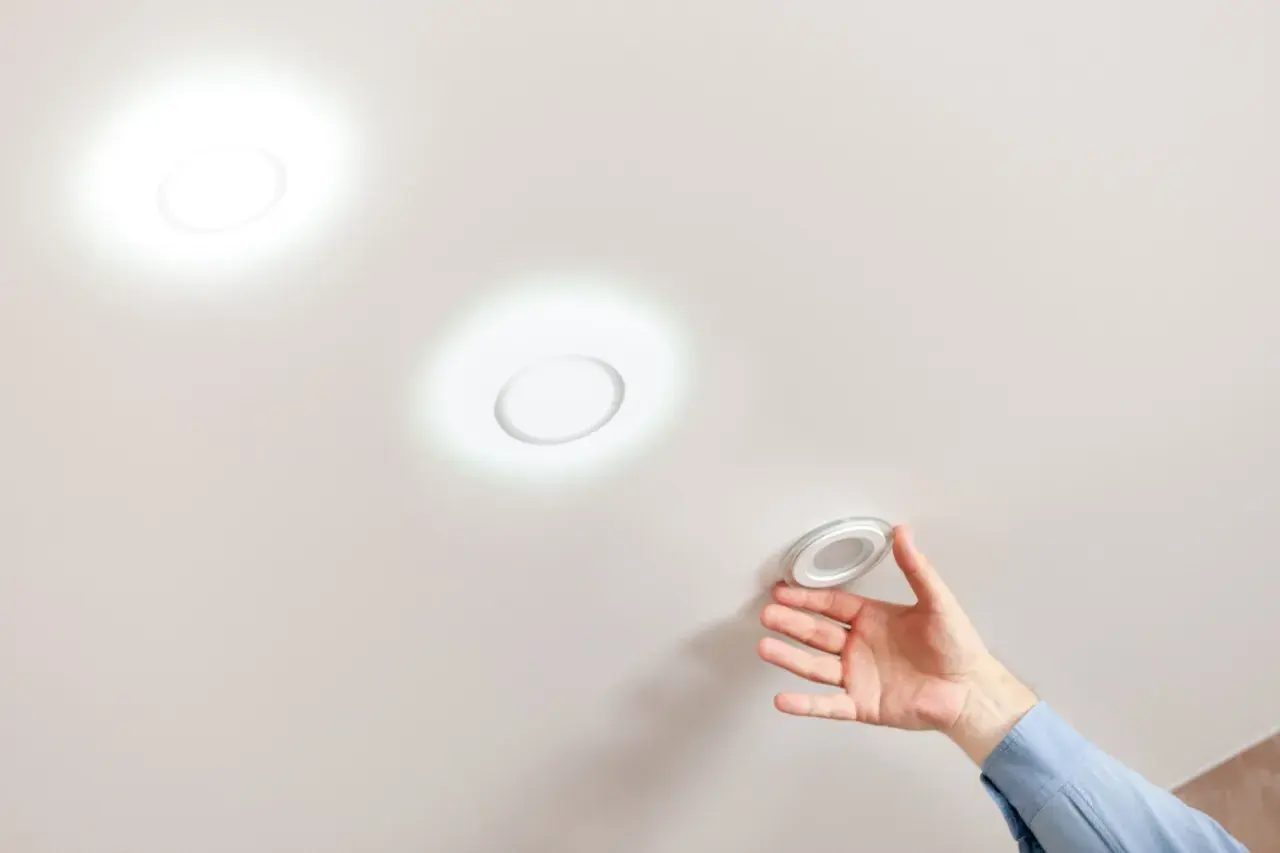 Mano instalando una lámpara techo redonda. Dos luces circulares ya iluminan el techo blanco.