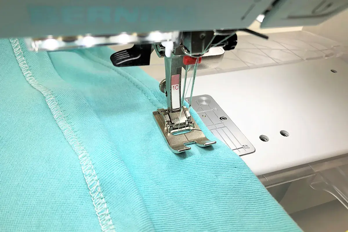 A double needle stitches a hem on turquoise fabric. Most sewing machines can use a double needle with the right setup.