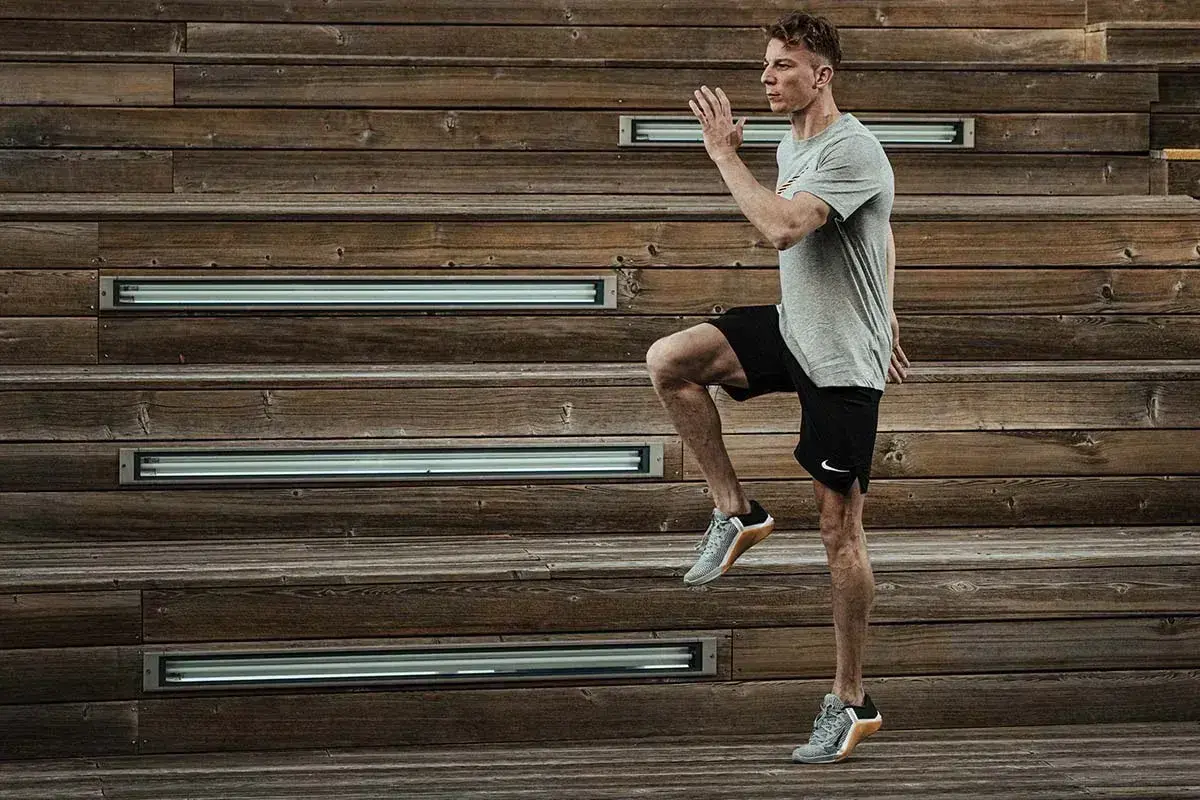 Mężczyzna w szarej koszulce i czarnych spodenkach wykonuje dynamiczne ćwiczenie, podnosząc kolano. Intensywny trening HIIT na drewnianych schodach.