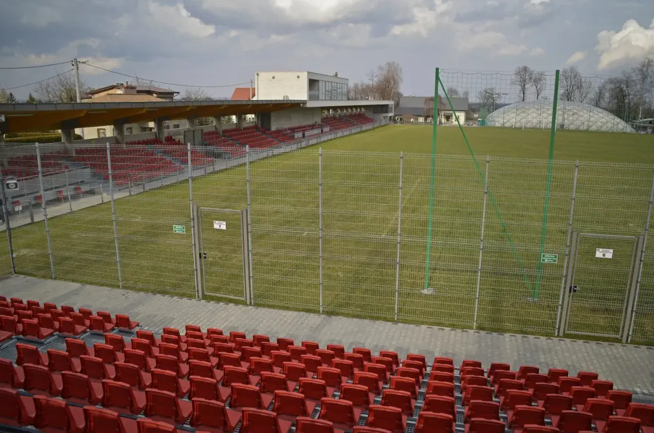 Puste trybuny i zielona murawa na stadionie w Niepołomicach. W tle budynek klubowy i balon nad boiskiem.