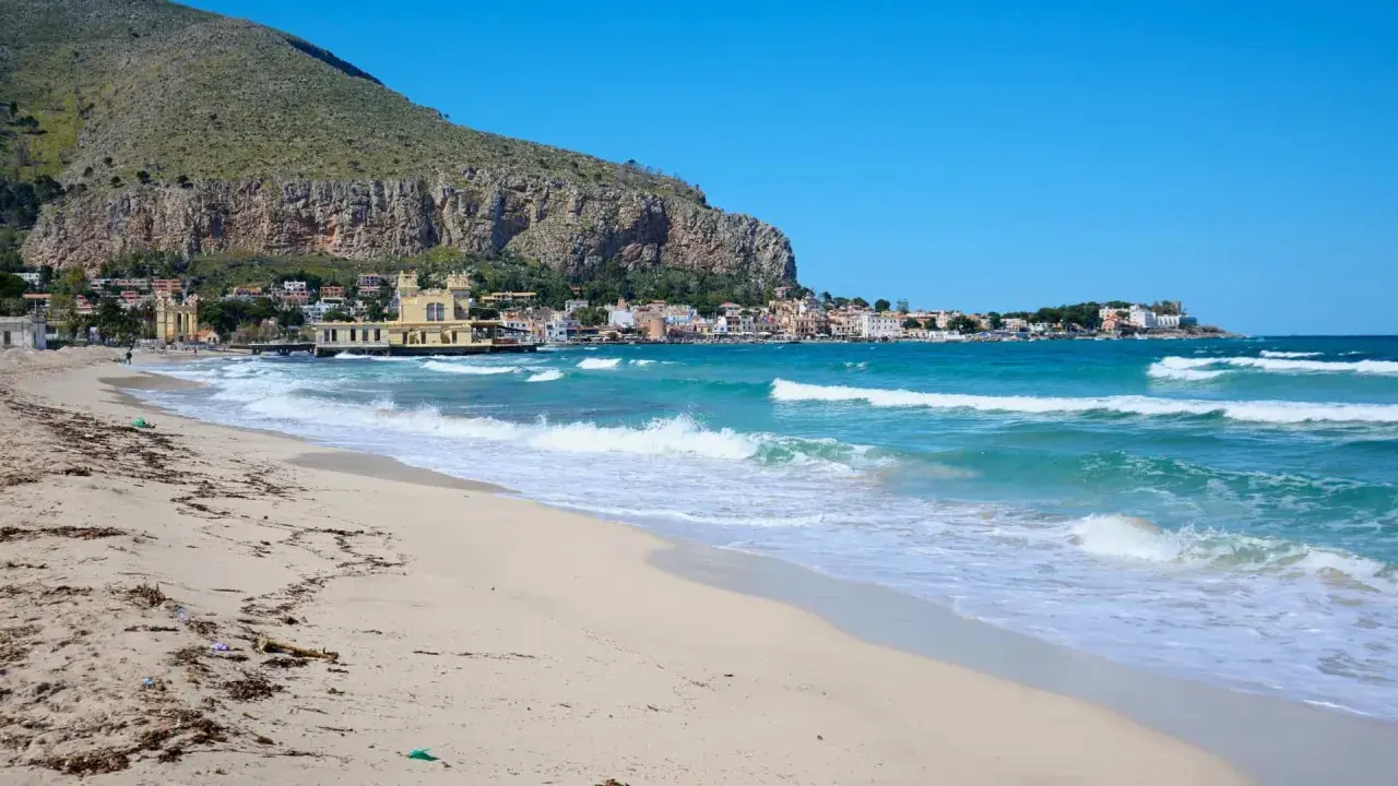 Spiaggia dorata e mare turchese in Sicilia, con un borgo colorato incastonato tra la montagna e l'acqua cristallina.